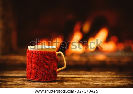 Cup of hot drink in front of warm fireplace. Holiday Christmas concept. Mug in red knitted mitten standing near fireside. Cozy relaxed magical atmosphere in a chalet.