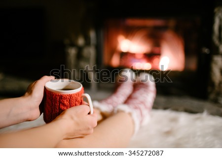 Woman holding a cup of tea by the Christmas fireplace. Woman relaxes by warm fire with a cup of hot drink and warming up her feet in woollen socks. Close up on feet. Winter, Christmas holidays concept