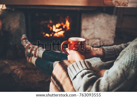 Feet in woollen socks by the Christmas fireplace. Woman relaxes by warm fire with a cup of hot drink and warming up her feet in woollen socks. Cozy atmosphere. Winter and Christmas holidays concept.
