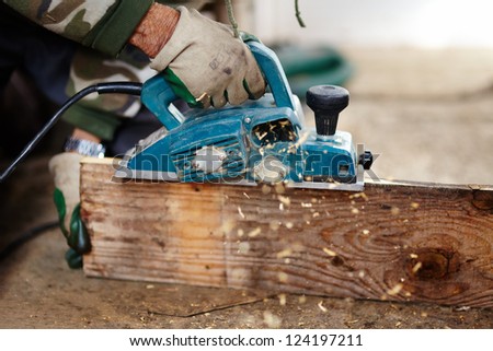 Man with protection gloves using an electric scraper to plane a plank Man with protection gloves using an electric scraper to plane a plank