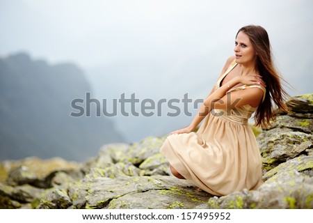 Beautiful young woman in a flowing nude dress sitting on the mountain rocks