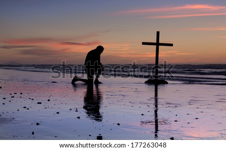 Man kneel at a cross on a beach at sunset.
