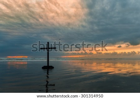 Black cross on a beach with a cloudy sunset in the background.