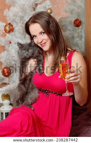 Beautiful young woman holding a glass of champagne and a black cat in front of a white Christmas tree.
