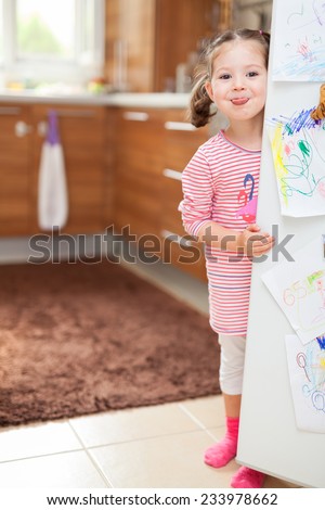 little cute girl sticking tongue out behind refrigerator door in kitchen.