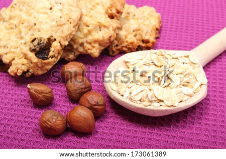 Oatmeal cookies and ingredients - oatmeal on wooden spoon and hazelnut lying on purple background