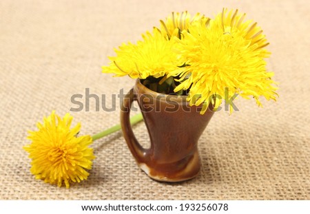 Bouquet of yellow fresh flowers of dandelion in brown vase standing on jute canvas