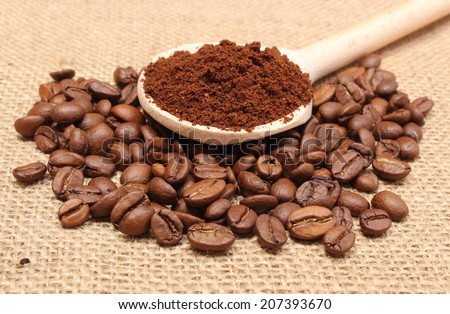 Ground coffee on wooden scoop lying on heap of coffee beans, coffee grains. Background texture of old jute