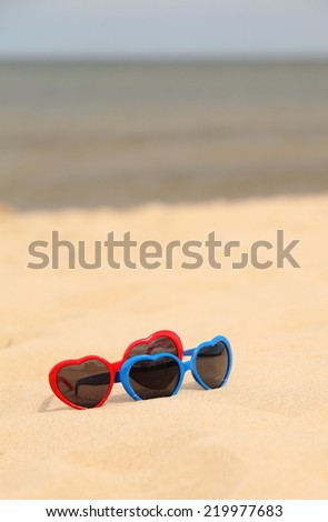 Blue and red funny sunglasses in the shape of a heart lying on the sand at the beach, summer time
