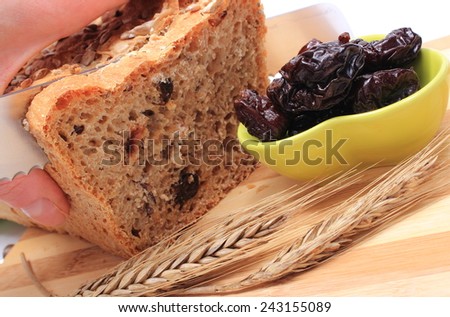 Slicing fresh baked wholemeal bread, heap of dried plums and ears of wheat lying on cutting board, concept for healthy eating