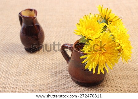 Bouquet of yellow fresh flowers of dandelion in brown vase and empty vase in background lying on jute canvas
