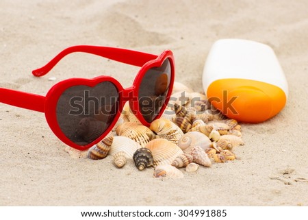 Heap of shells, red sunglasses in shape of heart and sun lotion on sand at the beach, seasonal concept, protection from sun