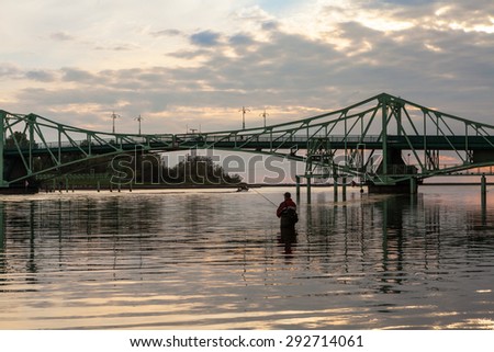 Fisherman stands alone in the water with fishing rod in front of the old steel structured bridge at sunset