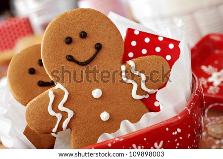 Smiling gingerbread men nestled in holiday dish with gift-wrapped surprise.  Baking ingredients and supplies (including baker\'s twine and rolling pin) in background.  Macro with extremely shallow dof.