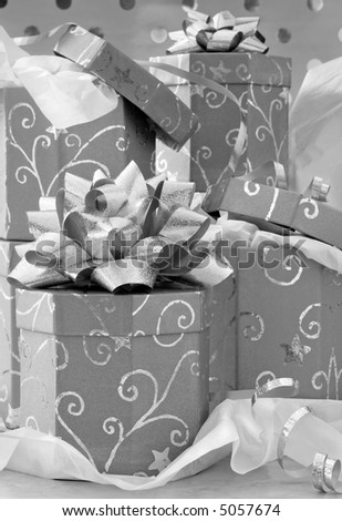 Black and white still life image of silver, ornate gift boxes with bows, ribbons, and tissue paper.  Silver wrapping paper in the background.
