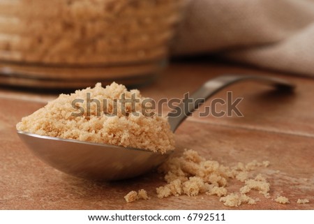 Macro still-life of serving spoon overflowing with brown sugar onto color coordinated porcelain tile. Glass canister of brown sugar and homespun cloth out of focus in the background.  Shallow dof