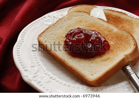 Freshly toasted bread with cherry preserves and serving knife on a decorative, antique plate.  Red damask table linen in the background.