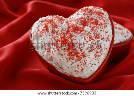 Individual sized, heart-shaped cakes with white frosting nestled in the soft folds of a red tablecloth.  Shallow dof.