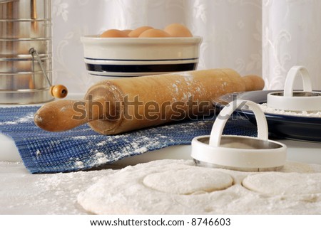 Vintage rolling pin with homemade biscuit dough.  Still-life with selective focus on flour sprinkled, rolling pin.  Bowl of eggs, flour sifter, and  lace curtains in soft focus in the background.
