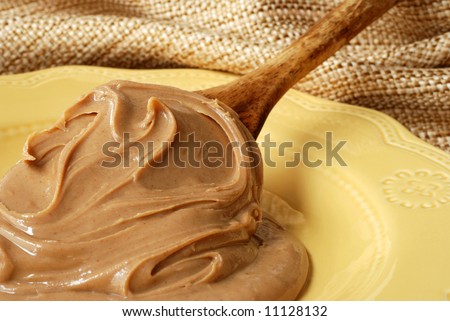 Creamy swirls of peanut butter on a vintage wooden spoon resting on a ceramic plate.  Natural, textured fabric in the background.  Shallow dof with focus on the peanut butter swirls.