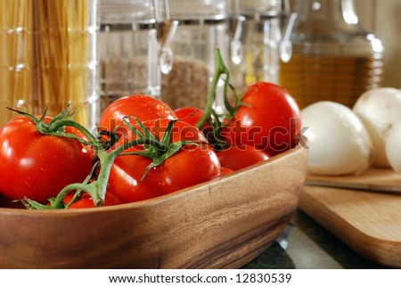 Freshly washed tomatoes in wooden bowl with onions, pasta, and glass canisters in the background.  Close-up with shallow dof and selective focus on closest tomato.