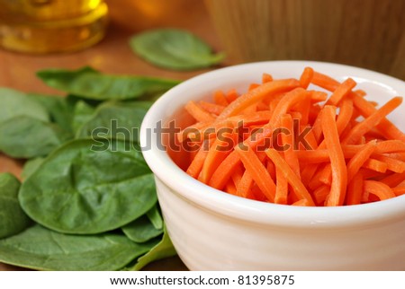 Freshly shredded carrots in small dish with baby spinach and salad oil in soft focus in background.  Macro with extremely shallow dof.