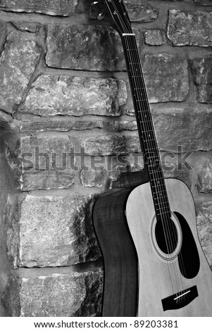 Rustic black and white image of a steel-stringed acoustic guitar leaning against a stone wall.