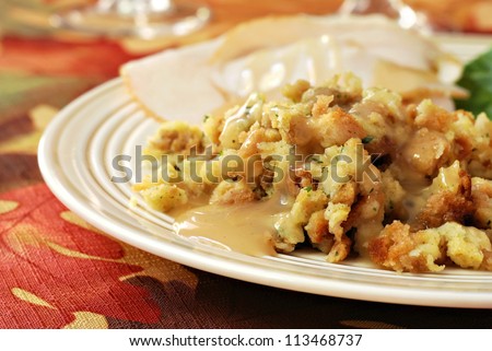 Homemade stuffing with gravy served on dinner plate (with turkey in background) on seasonal tablecloth for Thanksgiving.  Closeup with shallow dof.