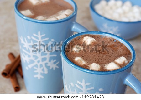 Snowflake mugs filled with hot chocolate and marshmallows on tile counter with cinnamon sticks along side. Matching bowl with marshmallows in soft focus in background. Closeup with shallow dof.