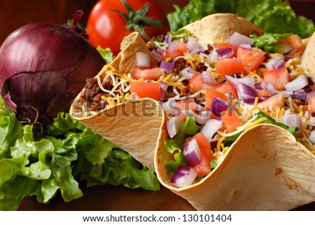 Taco Salad In Freshly Baked Flour Tortilla Bowl (Seasoned Ground Beef, Lettuce, Onions, Tomatoes, And Shredded Cheese) With Tomato, Onion, And Leaf Lettuce In Background. Macro With Shallow Dof.