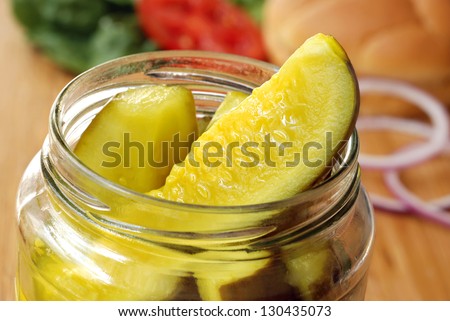 Dill pickle spears in open jar with sandwich ingredients in soft focus in background.  Macro with shallow dof.