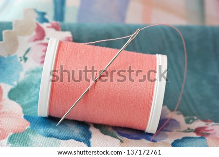 Threaded needle in spool of peach colored polyester thread with swatches of fabric.  Macro with shallow dof.  Selective focus on needle.