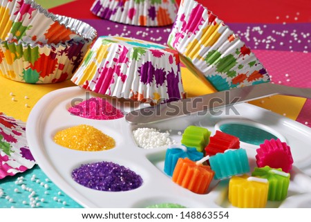 Colorful Still Life Of Cupcake Supplies With An Artist\'S Theme - Includes Wrappers With Splattered Paint Design And Palette Filled With Sugar Sprinkles And Candles.