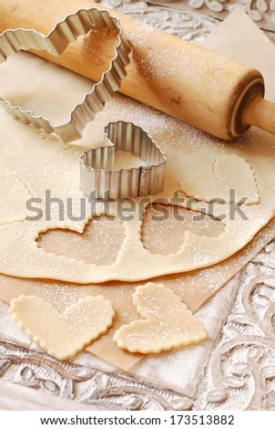 Heart shaped cookie cutters with dough and vintage rolling pin (dusted with flour) on rustic wood tray with carved design.