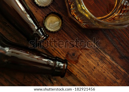 Top view of near empty beer glass with bottle caps and bottles on rustic wood background. Low key still life with directional natural lighting for effect. Selective focus on bottle caps and droplets.