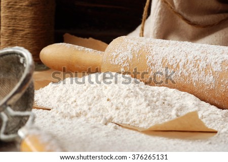 Baking still-life with flour heaped onto parchment paper, vintage wooden rolling pin, vintage sieve, and canvas flour sack with rope in background.  Closeup with shallow dof.  Selective focus on flour