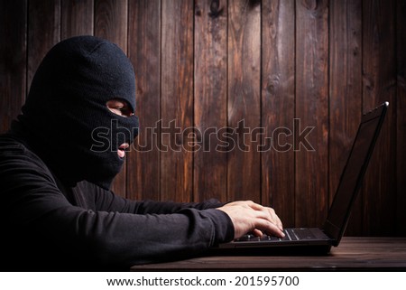 Hacker in a balaclava standing in the darkness furtively stealing data off a laptop computer on wooden background
