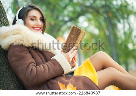 Young woman in autumn park reading book and listen music
