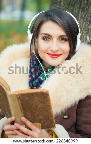 Young woman in autumn park reading book and listen music