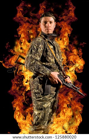 A soldier stands with a gun in his hand and safety glasses in a burning fire