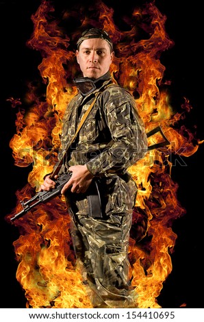 Soldier stands with a gun in his hand and safety glasses in a burning fire