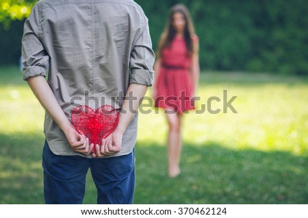 Young man standing in front of a young woman holding a heart behind his back. Saint Valentine\'s day.