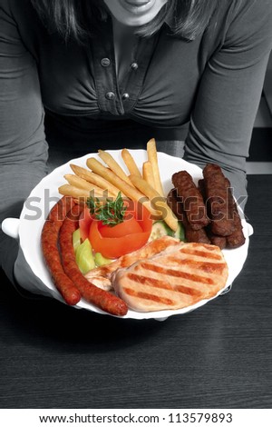 woman in gray scale technique offering colorful grill with french fries and vegetables on a white plate. Accent is on the food, and for that reason, small noise added on black and white background.