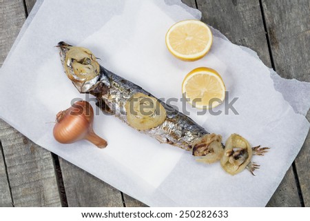 Top view of baked mackerel fish on white paper with baked slices of onion on top; with lemon and whole onion. Wooden background.