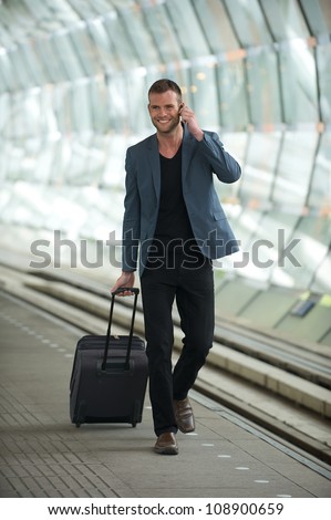 Happy smiling young business man wearing casual suit and walking in the train station with a phone and suitcase in hand