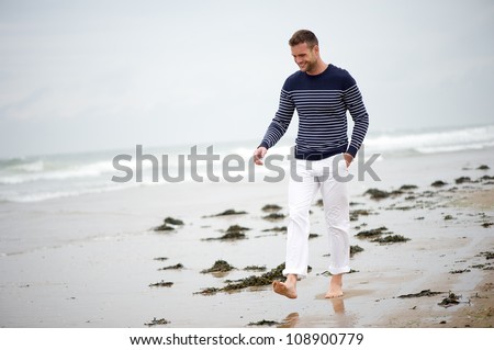 Handsome young man walking on the sand at the beach. He is barefoot and smiling.