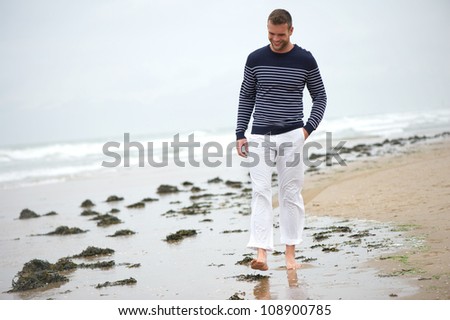 Handsome young man walking alone and smiling on the beach. He is barefoot in the sand.