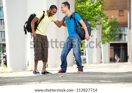 Two young male students walking and laughing outdoors on college campus. Full length portrait of a young African American male student with Caucasian student