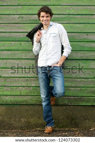 Handsome young man leaning against a green wooden wall outdoor. He is holding his jacket and hand in pocket. One leg up