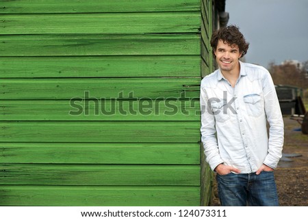 Happy male model leaning against green wooden wall outdoor. Hands in pockets and smile expression on his face